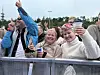 Jublende publikummere står bak et metallgjerde og holder drikke i hendene.