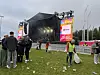 Utendørs festivalscene med publikum, vakter, gjerder og søppel på gresset.