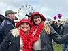 To smilende kvinner med røde hatter og blomsterkranser foran et pariserhjul og en folkemengde.