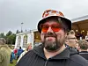 Skjeggete mann med solbriller og hatt smiler foran et utendørsarrangement.
