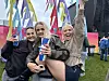 Tre kvinner smiler på en gressplen foran fargerike flagg og festivalpublikum.