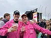 To menn i rosa klær holder drikke foran en utendørsscene med festivalpublikum rundt seg.