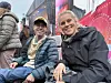 To personer smiler mot kameraet i en travel bygate med fargerike skilt i bakgrunnen.