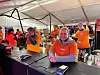 Smilende medarbeidere i oransje klær står bak en serveringsdisk med drikkevarer og utstyr.