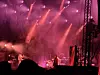 Musikere spiller på en scene under rødt og rosa lys, med scenestillas og publikum i forgrunnen.