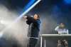 Artist på scenen peker mot publikum, mens en musiker står bak et miksebord i blått lys og scenetåke.
