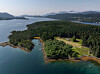 Flyfoto av en skogkledd øy med en smal vik, små bygninger og fjellkledd landskap i bakgrunnen.