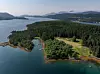 Flyfoto av en skogkledd øy med en smal vik, små bygninger og fjellkledd landskap i bakgrunnen.