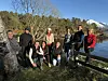En gruppe voksne står og sitter samlet utendørs ved vannet, med skog og fjell i bakgrunnen.
