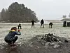 En gruppe personer trener på et snødekt område ved sjøen, mens en person sitter på huk i forgrunnen.