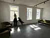 Nesten tomt rom med sofaer, tre vinduer og to personer ved et vindu.