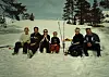 Seks personer sitter og står samlet i snøen med ski og staver.