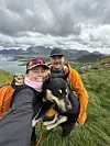 To turgåere i fargerike klær holder en svart hund foran et fjellandskap under skyet himmel.
