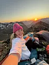 En kvinne i rosa lue tar en selfie blant turutstyr i fjellet mens solen går ned.