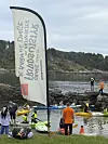 Mennesker i redningsvester bruker kajakker og SUP-brett ved en strand. Et stort banner står i forgrunnen.