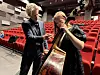 To personer står i en konsertsal med en kontrabass foran rader med røde stoler.