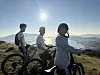 Tre syklister står på en høyde i solskinn med utsikt over fjord og fjell.