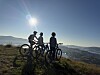 Tre syklister står med sykler på en fjellrygg i motlys over et fjordlandskap.