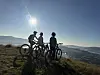 Tre syklister står med sykler på en fjellrygg i motlys over et fjordlandskap.