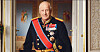 Eldre mann i mørk militæruniform med medaljer og blått bånd i et utsmykket rom.