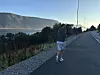 En løper i shorts og blå genser beveger seg på en vei med fjord og fjell i bakgrunnen.