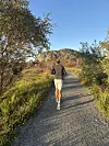 En person løper på en grussti omgitt av trær, med fjell og blå himmel i bakgrunnen.