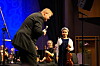 Dirigent bøyer seg mot en ung cellist på scenen, omgitt av musikere og instrumenter.