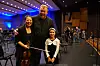 Et barn med fiolin står mellom to voksne i en konsertsal med orkesterstoler og publikum i bakgrunnen.