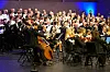 Dirigent leder et stort kor og orkester på en opplyst konsertscene.