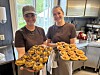 To kvinner står på et kjøkken og holder brett med nybakte kanelsnurrer og annen søtbakst.