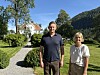 To personer står på en grussti i en hage foran en hvit villa med fjell i bakgrunnen.