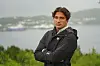 Mann i svart jakke står med armene i kors foran et fjordlandskap.