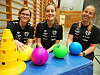 Tre unge kvinner i svarte treningsskjorter står bak fargerike baller og kjegler i en gymsal.
