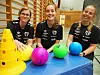 Tre unge kvinner i svarte treningsskjorter står bak fargerike baller og kjegler i en gymsal.
