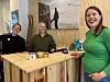 Tre smilende personer står i et trekledd lokale ved en disk med kaffekopper.