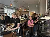 Travelt kaféinteriør med ansatte bak disken og flere kunder ved bardisken.