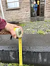 En hånd holder et målebånd mot en høy fortauskant utenfor en steinbygning.