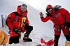To personer i rødt vinterutstyr står på snøen ved et fjell, med telt og utstyr rundt seg.