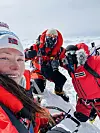 Fjellklatrere i rødt utstyr står i snødekt terreng under en overskyet himmel.