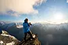 Vandrer i blå jakke står på en steinete fjellkam foran snødekte fjell og lav sol.