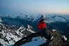 Person i rød jakke sitter på en snødekt fjellrygg foran en bred fjellkjede.