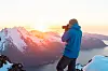 Person i blå jakke fotograferer fjell og fjord i varmt solnedgangslys.
