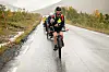 Syklist med hjelm og turutstyr sykler på en våt vei i et høstpreget fjellandskap.