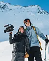To personer i skiklær står i snøen foran et fjell; den ene holder et videokamera.