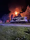 Et bolighus står i brann bak en stor campingvogn, med flammer og røyk mot nattehimmelen.