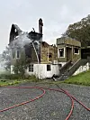Et utbrent trehus med røyk fra taket og flere brannslanger på bakken.