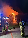 To brannmannskaper står ved en brannbil mens flammer brenner bak trærne om natten.