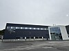 Moderne mørkt næringsbygg med glassinngang og parkeringsplass i overskyet vær.