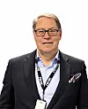 Portrett av en smilende mann i mørk dress, briller og navneskilt mot hvit bakgrunn.