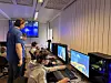 Barn og unge spiller dataspill ved flere skjermer i et rom, mens en voksen står ved siden av.